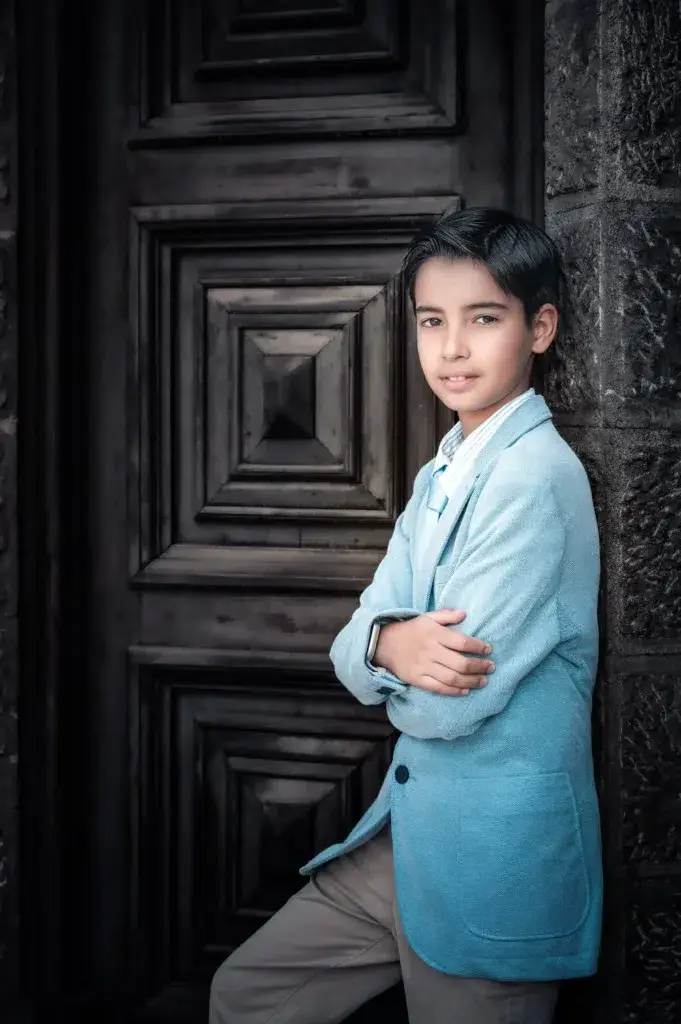 Niño con traje azul junto a una puerta de madera en su sesión de comunión.